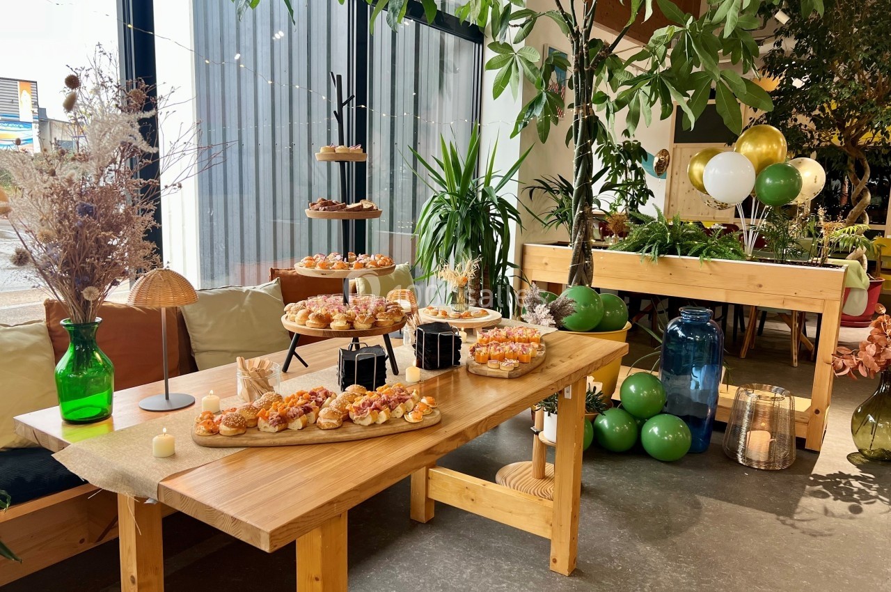 Table en bois décorée avec des plantes, des ballons et des plateaux de bouchées apéritives dans un espace lumineux.