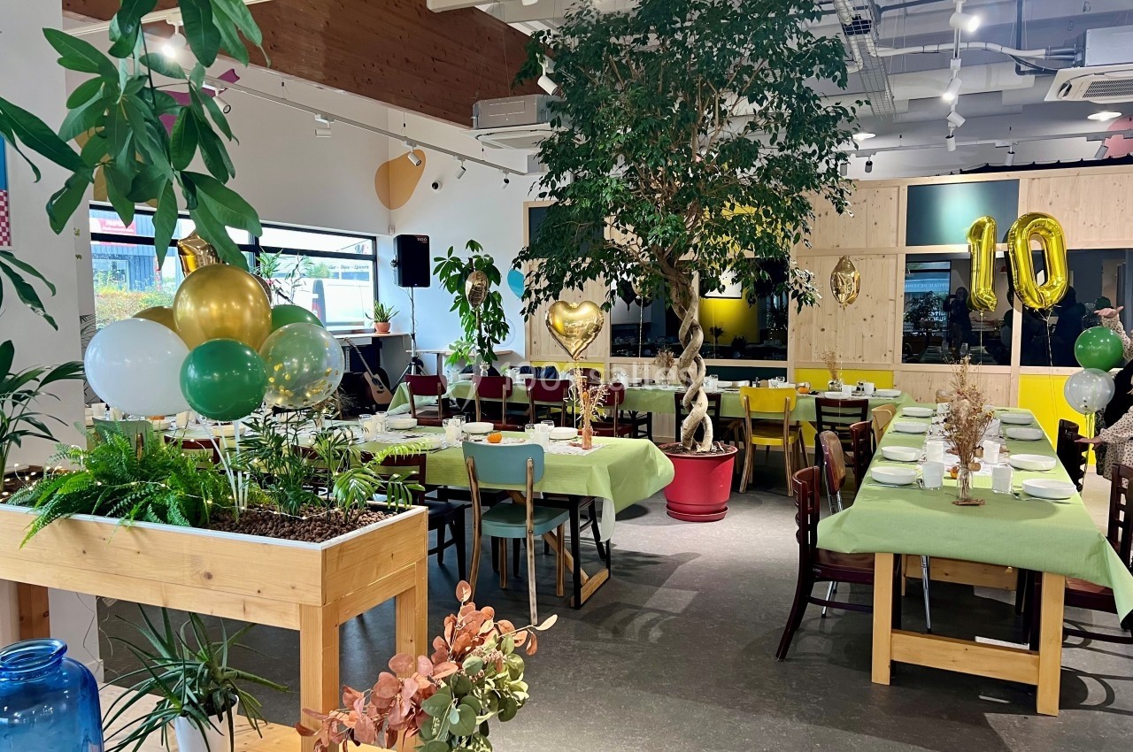Salle décorée pour une fête avec des ballons dorés, des tables dressées et des plantes en arrière-plan.