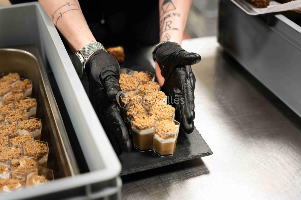 Personne disposant des verrines individuelles garnies de crumble sur un plateau dans une cuisine professionnelle.