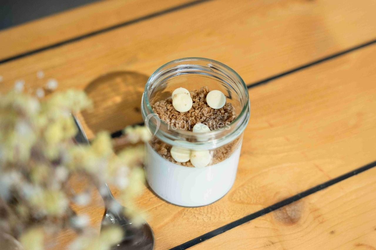 Dessert en pot avec une base blanche, surmonté de crumble et de pépites de chocolat blanc, posé sur une table en bois.