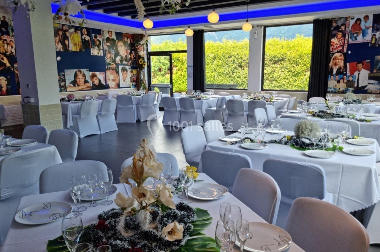 Salle de réception décorée avec des tables dressées, nappes blanches et compositions florales, murs ornés de photos.
