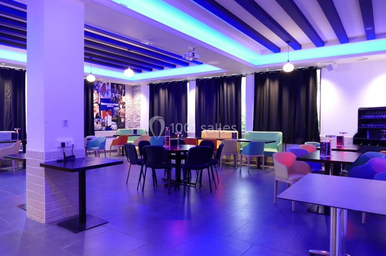 Salle moderne avec tables et chaises colorées, éclairage bleu au plafond et rideaux noirs couvrant les fenêtres.