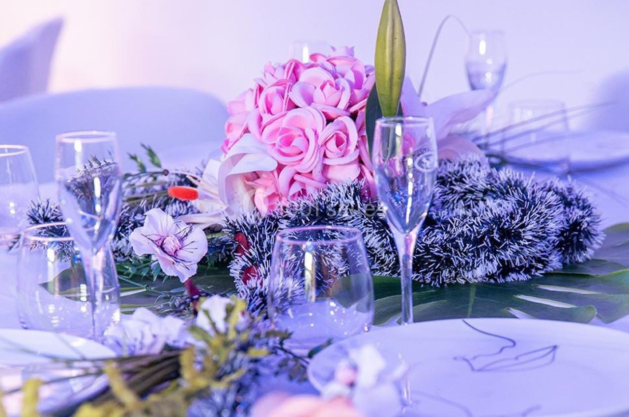 Centre de table avec bouquet de roses roses, feuillage décoratif et verres disposés sur une nappe blanche.