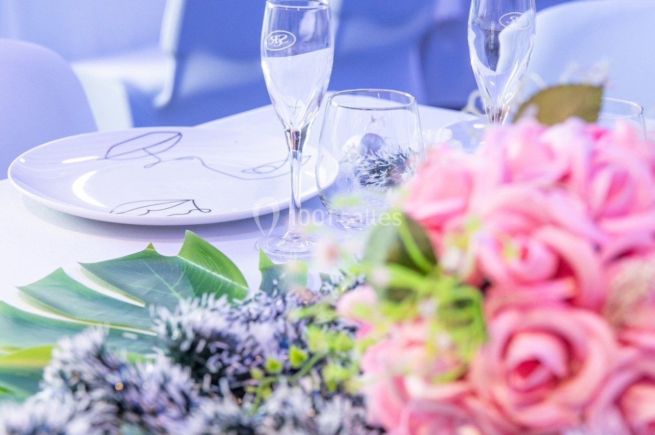 Table dressée avec assiettes blanches, verres à pied, bouquet de roses roses et feuillage décoratif.