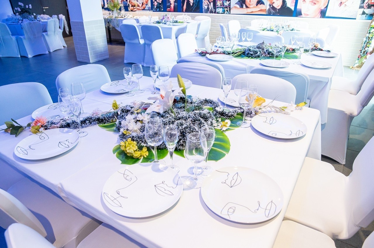 Table décorée pour un événement, avec nappes blanches, assiettes, verres et un centre de table floral coloré.