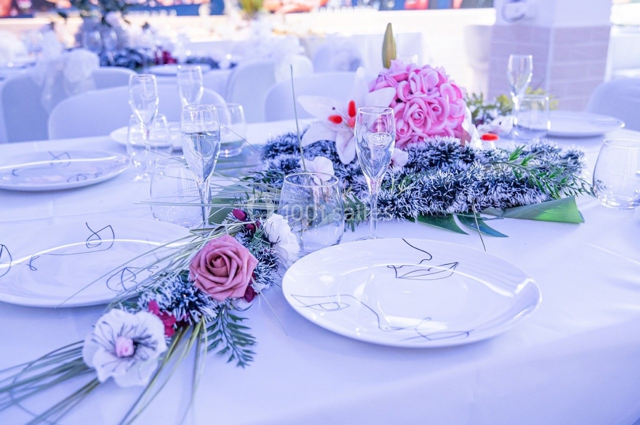 Table dressée avec vaisselle blanche, verres à pied et décoration florale composée de roses et de feuillage.