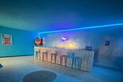 Bar rétro avec tabourets colorés, mur bleu, éclairage néon et jukebox dans un décor vintage.