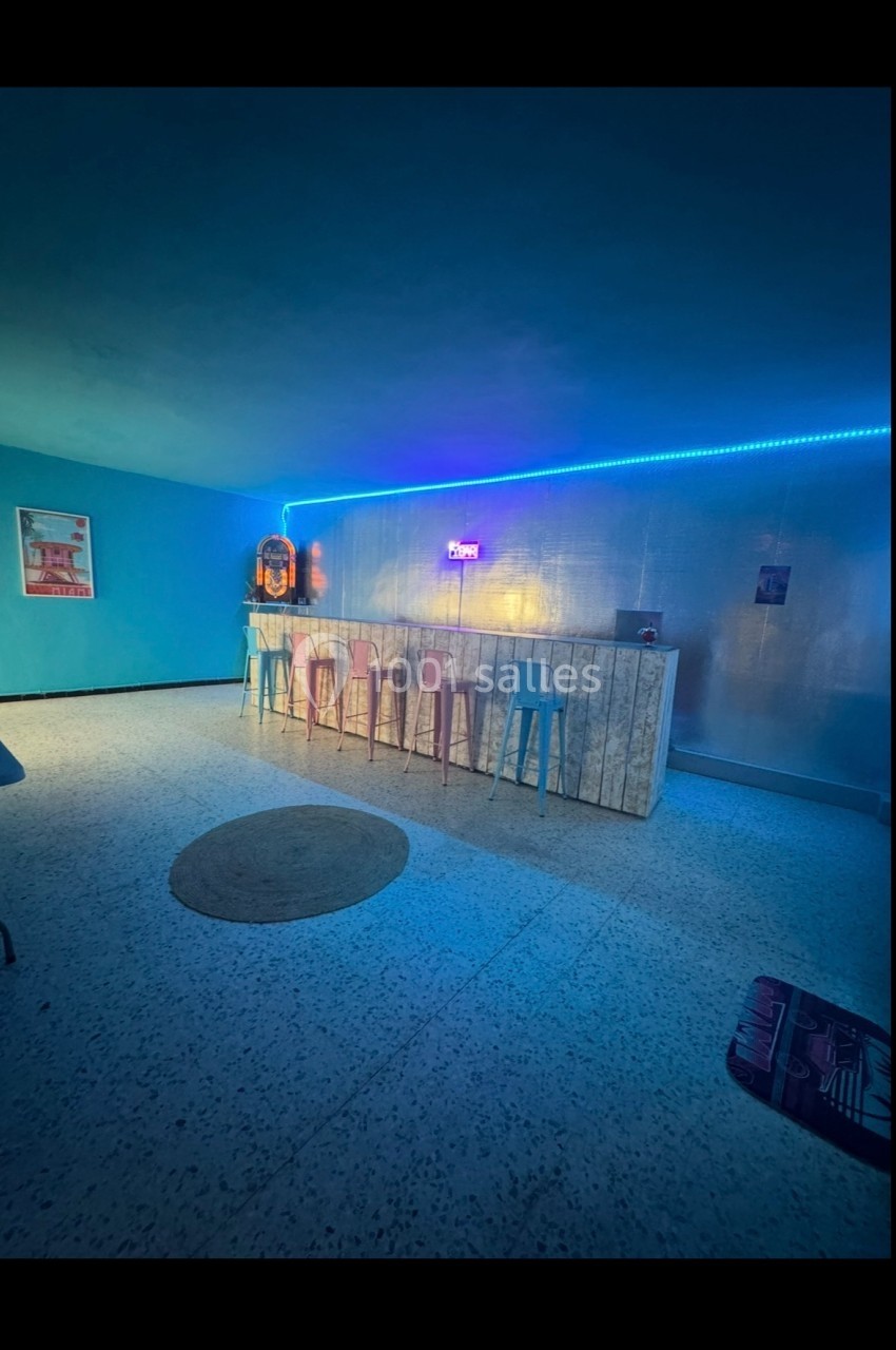Salle rétro avec un bar en bois clair, tabourets colorés, néons au mur et ambiance lumineuse bleutée.
