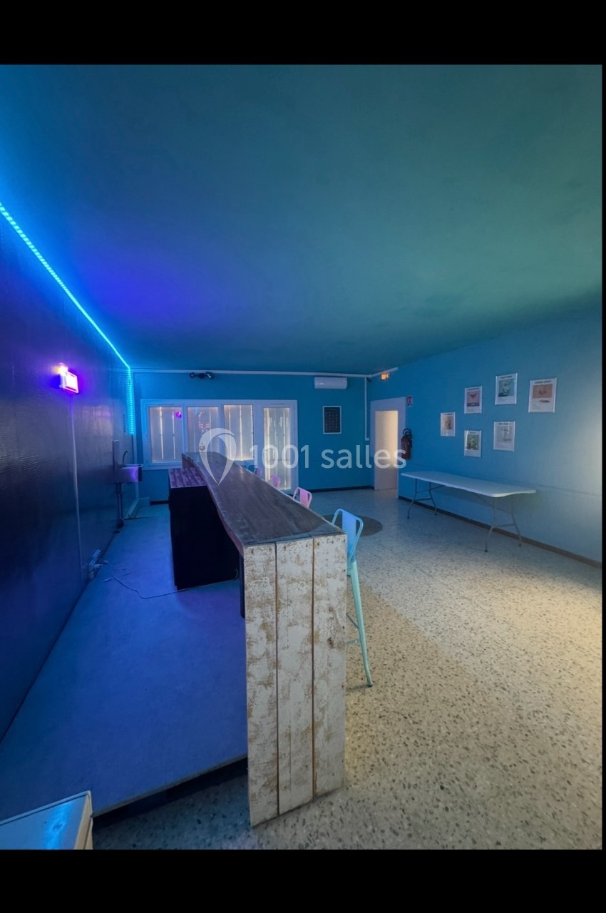 Salle intérieure avec murs bleus, table en bois au centre, chaises colorées et éclairage LED violet.