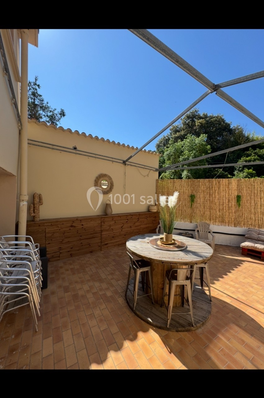 Terrasse ensoleillée avec table ronde en bois, chaises, plantes décoratives et paroi en bambou sous un ciel dégagé.