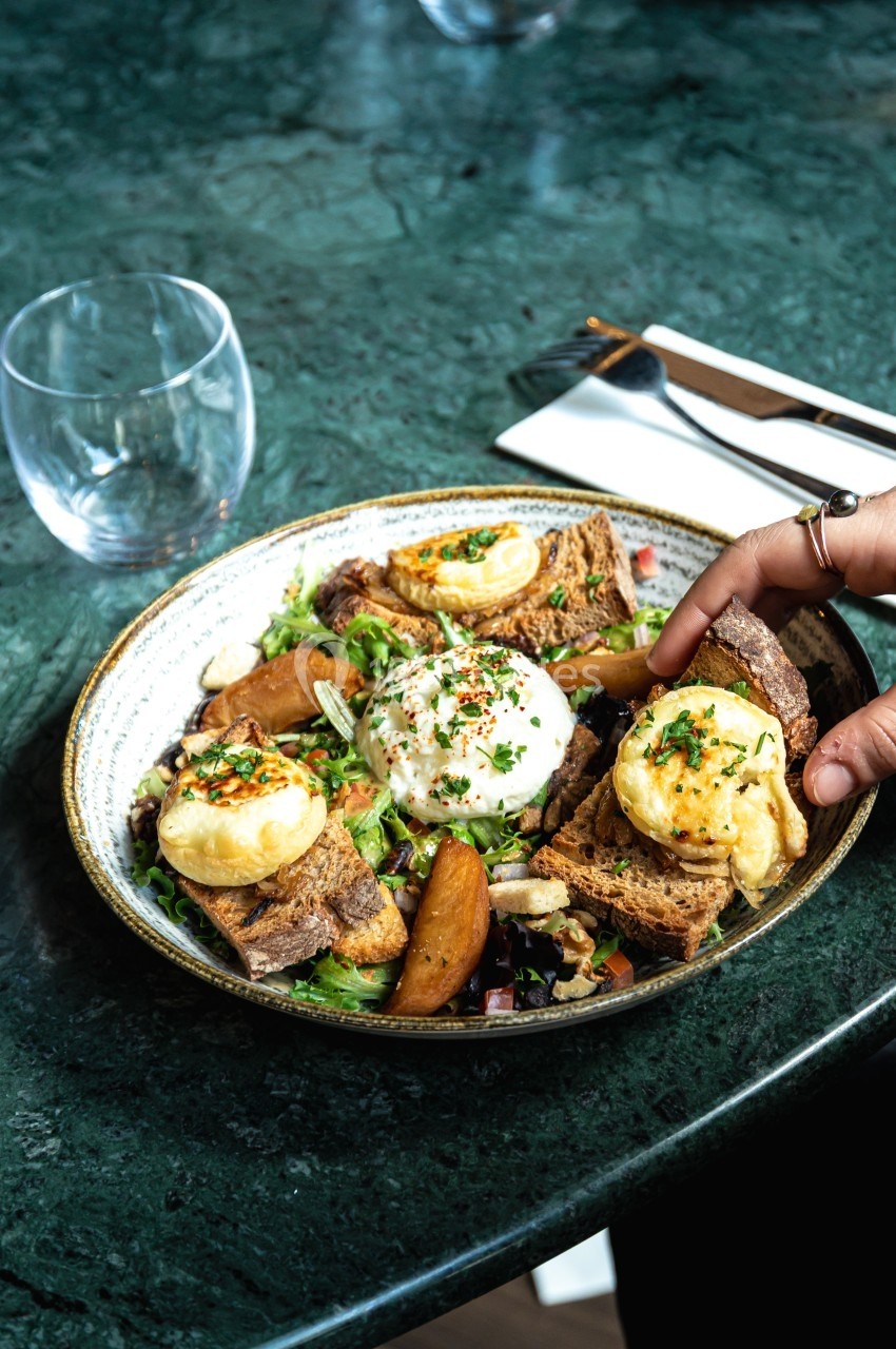 Assiette de salade garnie de fromage chaud sur pain grillé, pommes de terre rôties et œuf poché, servie sur une table verte.