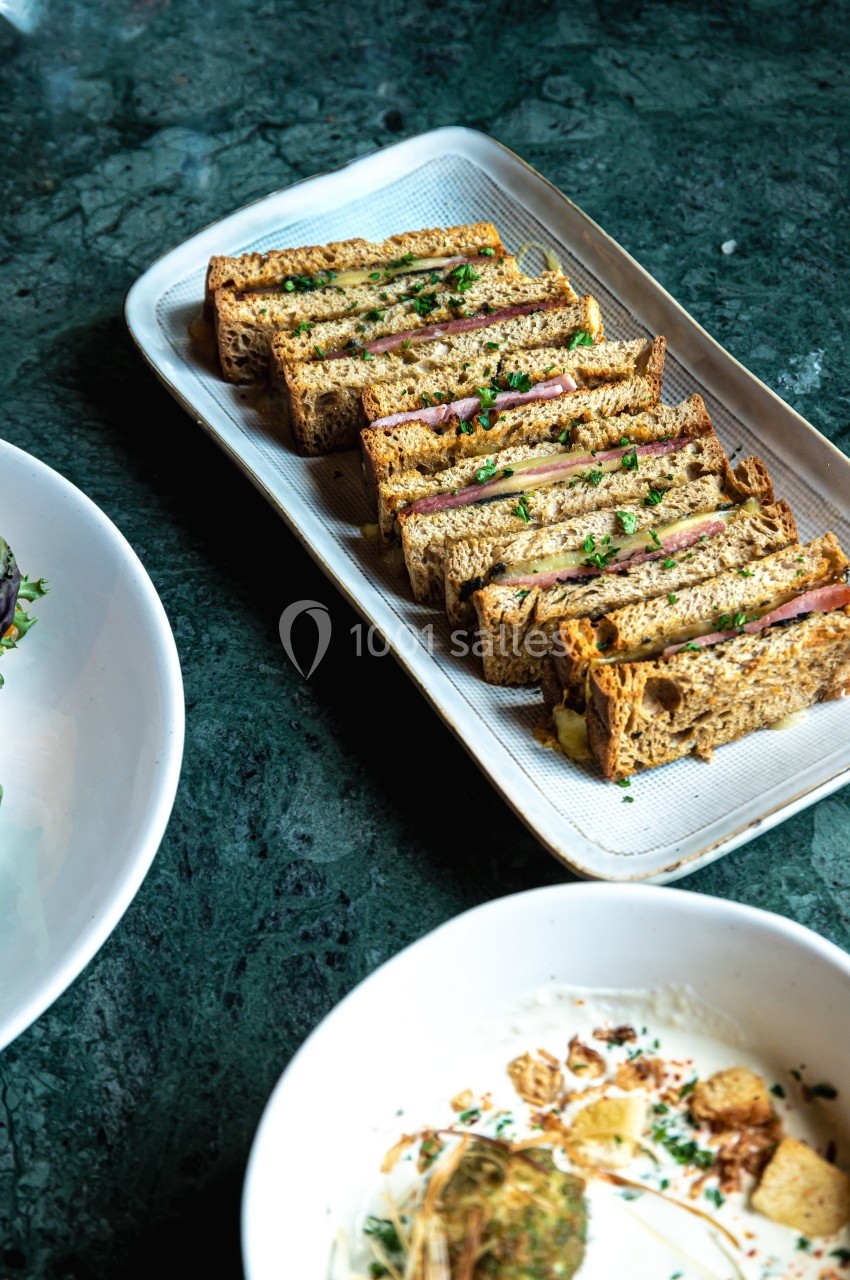 Assiette de sandwichs grillés garnis, présentés en tranches sur une table en marbre vert, accompagnés de plats blancs.