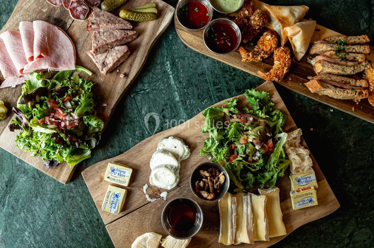 Assortiment de charcuteries, fromages, salades et condiments disposés sur des planches en bois sur une table sombre.