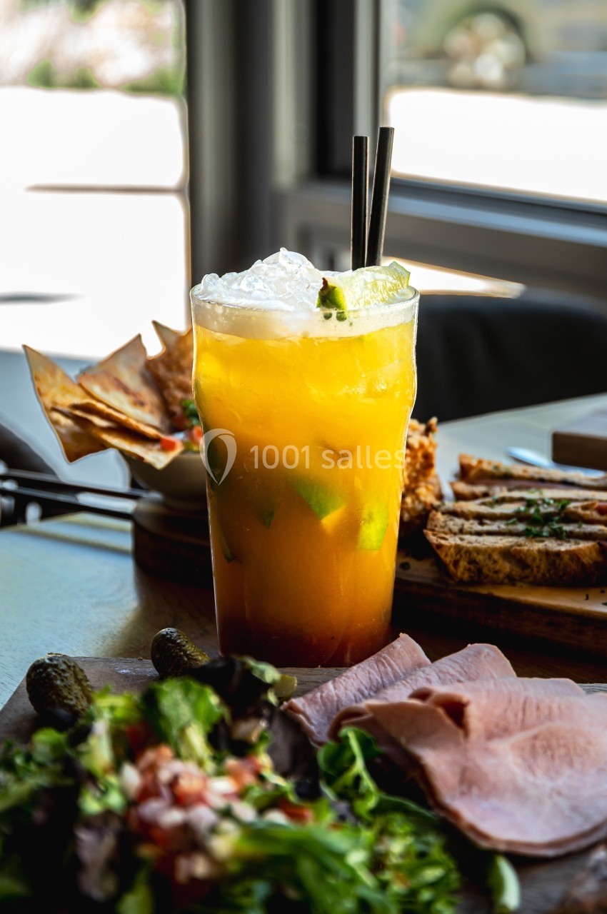 Verre de cocktail orange garni de glace et citron, entouré de charcuterie, salade et pain sur une table.