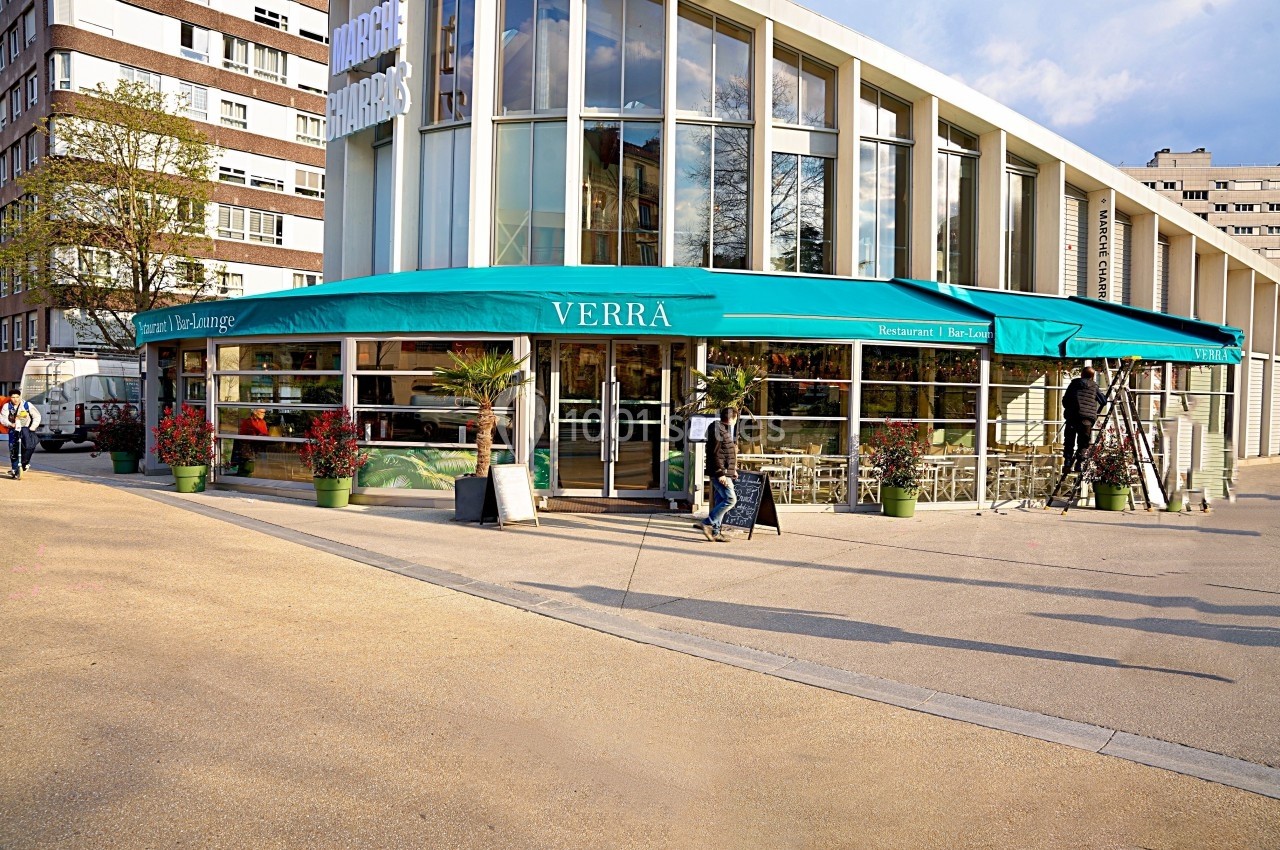 Façade d'un restaurant avec terrasse, enseigne ’Verrà’, entouré de plantes et situé près d'immeubles modernes.