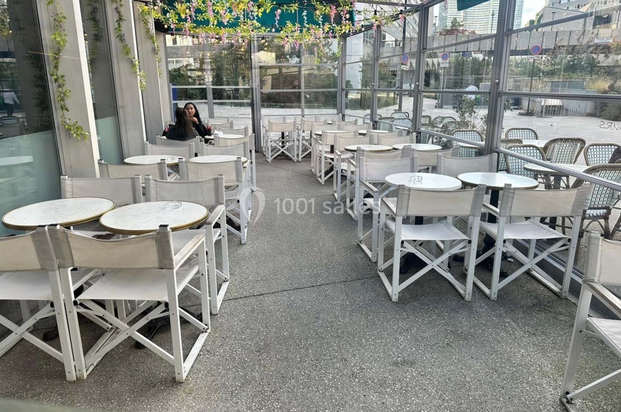 Terrasse extérieure avec tables et chaises blanches, partiellement couverte par une pergola ornée de plantes suspendues.