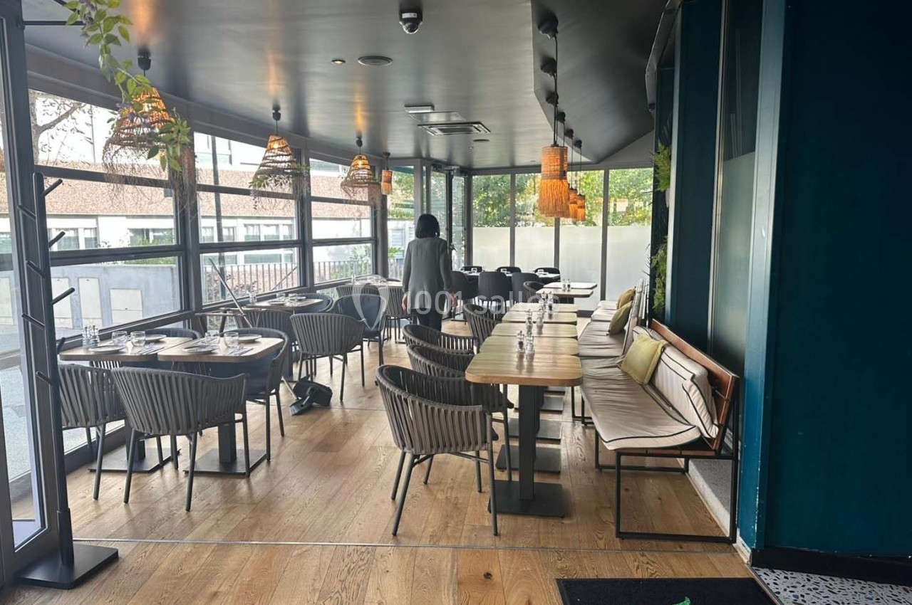 Salle de restaurant lumineuse avec grandes baies vitrées, mobilier en bois et rotin, et décoration végétale.