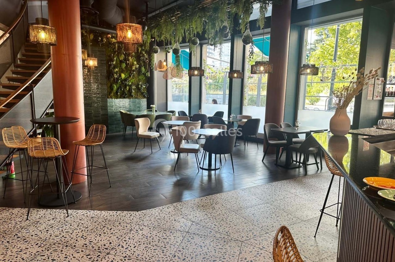Salle de restaurant moderne avec tables, chaises variées, plantes suspendues et grandes baies vitrées donnant sur l…