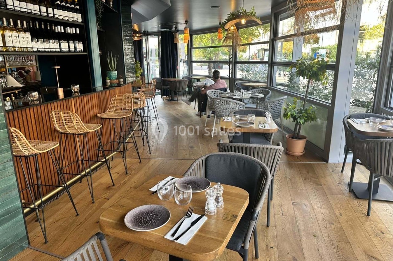 Salle de restaurant lumineuse avec tables dressées, bar en bois, chaises en rotin et grandes baies vitrées donnant sur l…