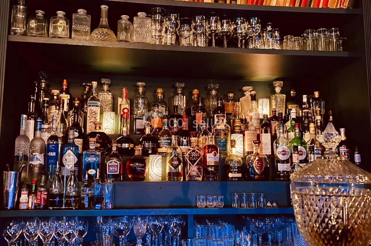 Étagères remplies de bouteilles d'alcool, verres et livres, derrière un comptoir en bois dans un bar.