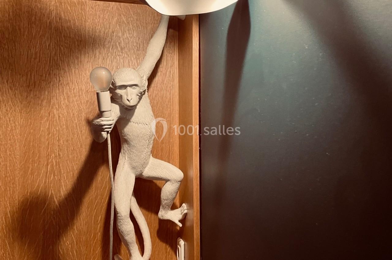 Singe sculpté tenant une ampoule, suspendu à une lampe blanche dans un décor en bois et mur sombre.
