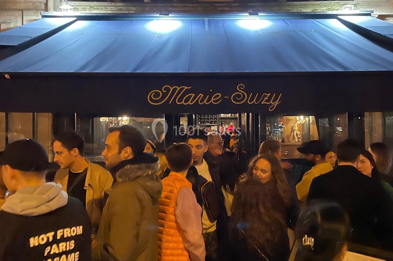 Groupe de personnes rassemblées devant un établissement nommé ’Marie-Suzy’ avec une façade éclairée en soirée.