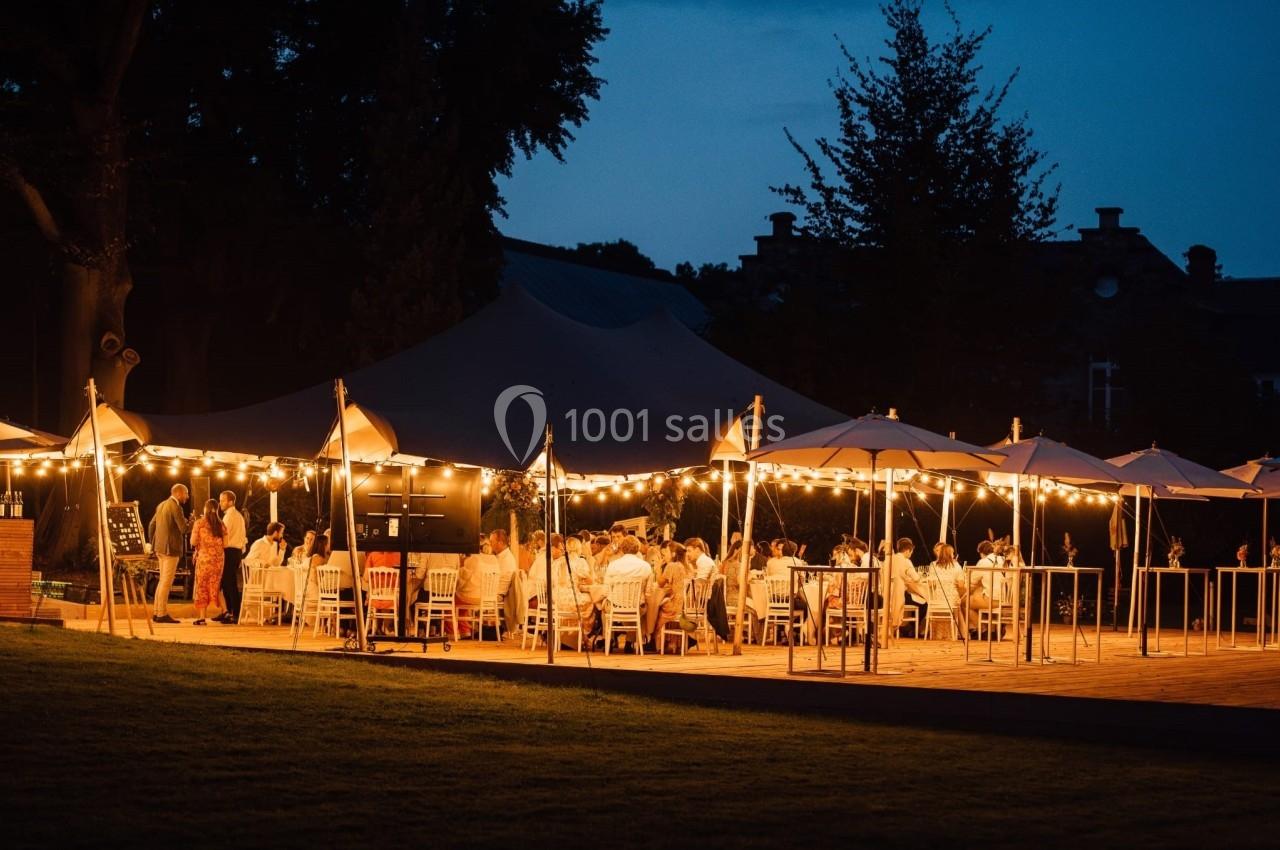 Des invités dînent en extérieur sous des guirlandes lumineuses et des parasols, dans une ambiance nocturne festive.