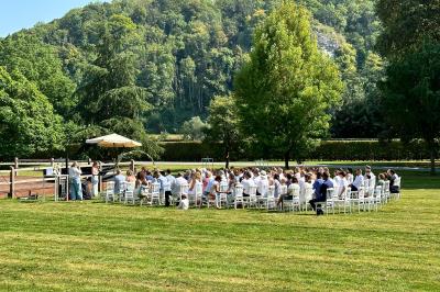 Groupe de personnes assises en extérieur lors d'une cérémonie, entourées d'arbres et de fleurs sous un ciel partiellement…