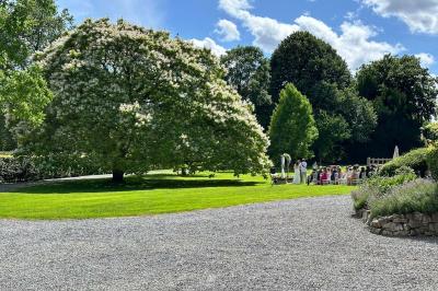 Groupe de personnes assises en extérieur lors d'une cérémonie, entourées d'arbres et de fleurs sous un ciel partiellement…