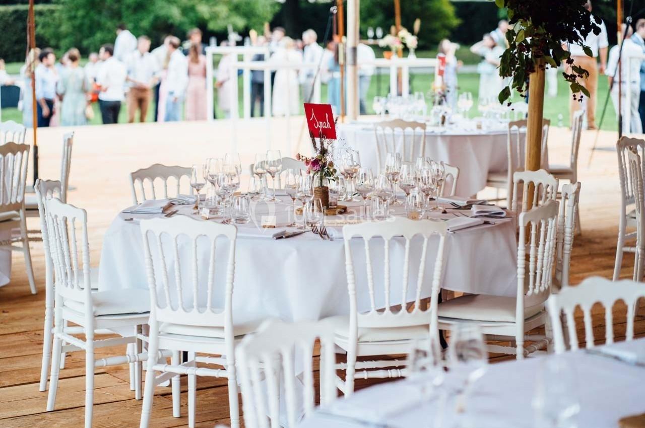 Tables rondes dressées avec nappes blanches, chaises en bois blanc et décorations florales, lors d'un événement en extérieur.
