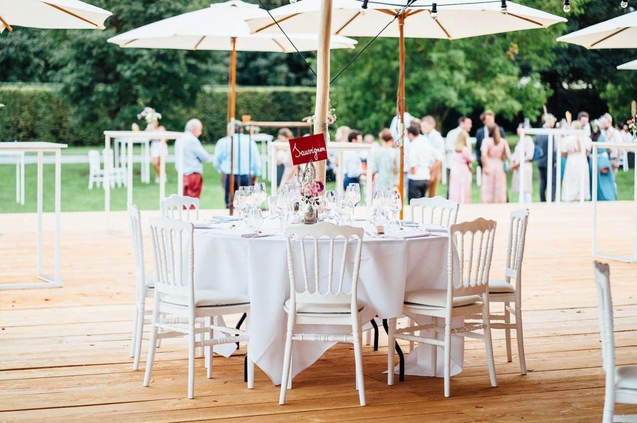 Table ronde dressée avec nappes blanches et chaises, située en extérieur sous des parasols, avec des invités en arrière-plan.