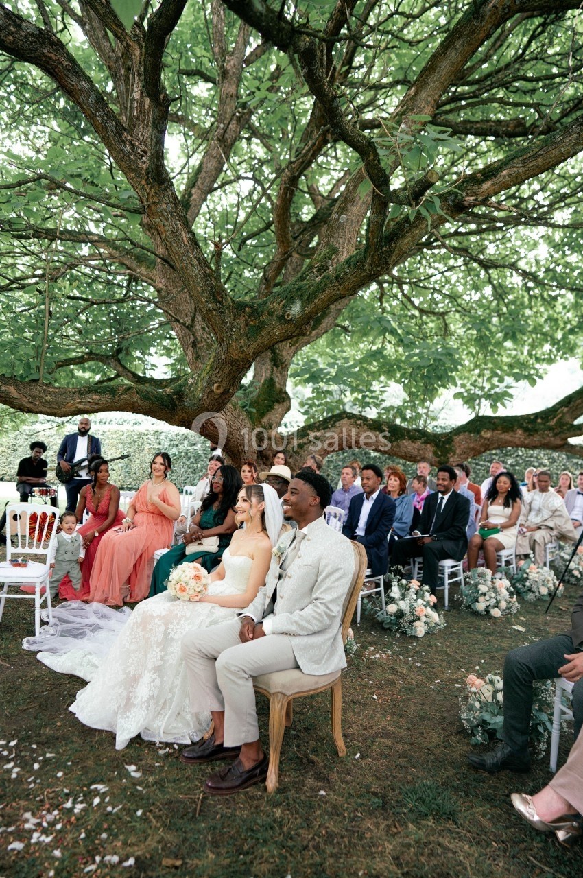 Un couple assis sous un grand arbre, entouré d'invités lors d'une cérémonie de mariage en plein air.
