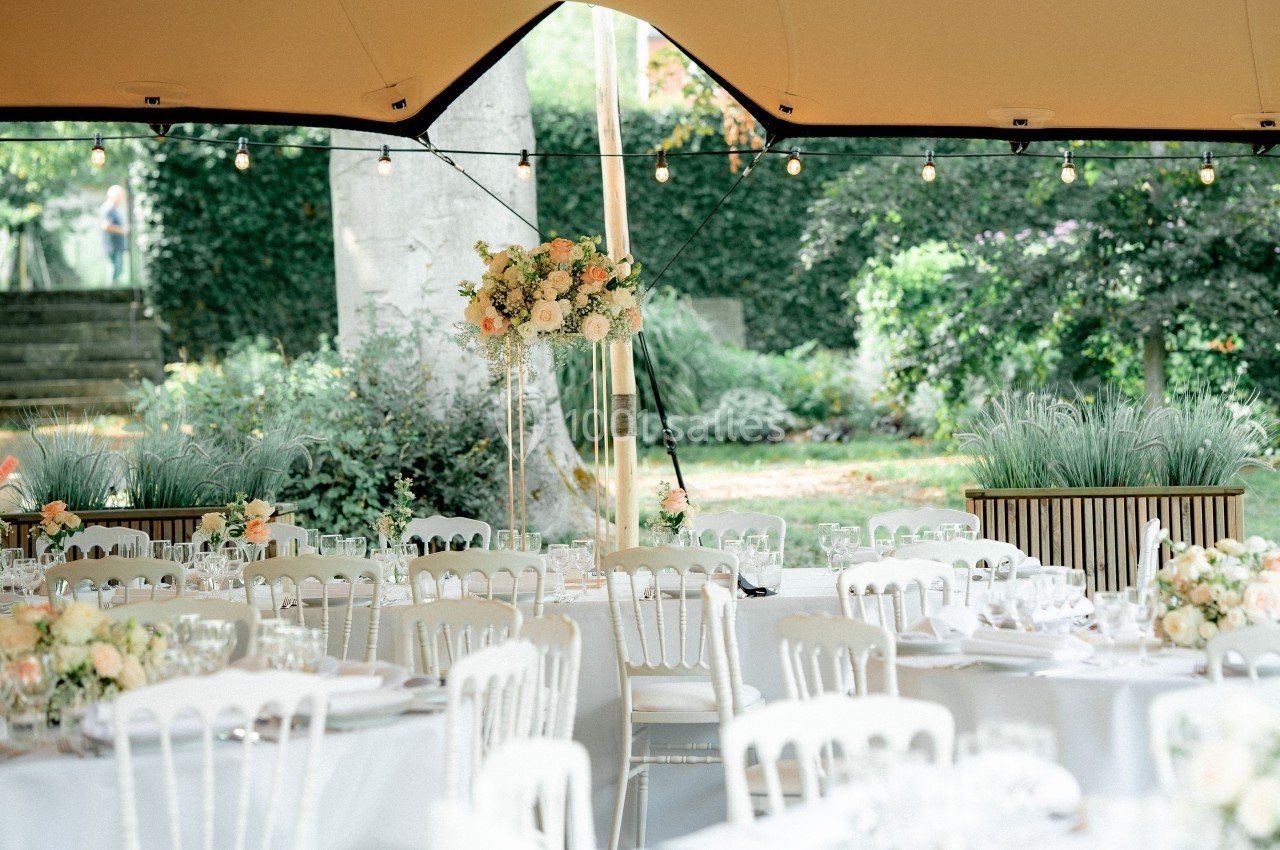 Salle de réception en extérieur avec tables dressées, chaises blanches et décoration florale sous une tente éclairée.