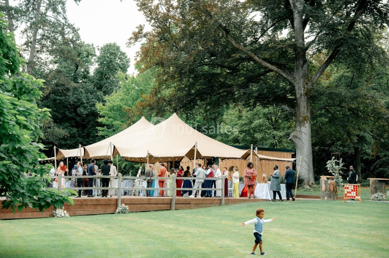 Tente beige installée dans un jardin, entourée d'invités debout, avec un enfant courant sur la pelouse au premier plan.