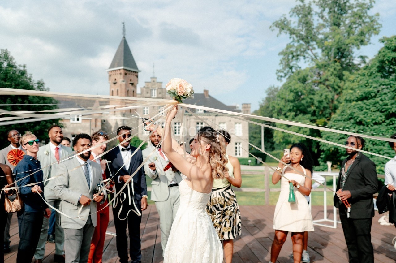 Une mariée lance son bouquet entourée d'invités souriants dans un jardin près d'un bâtiment historique.