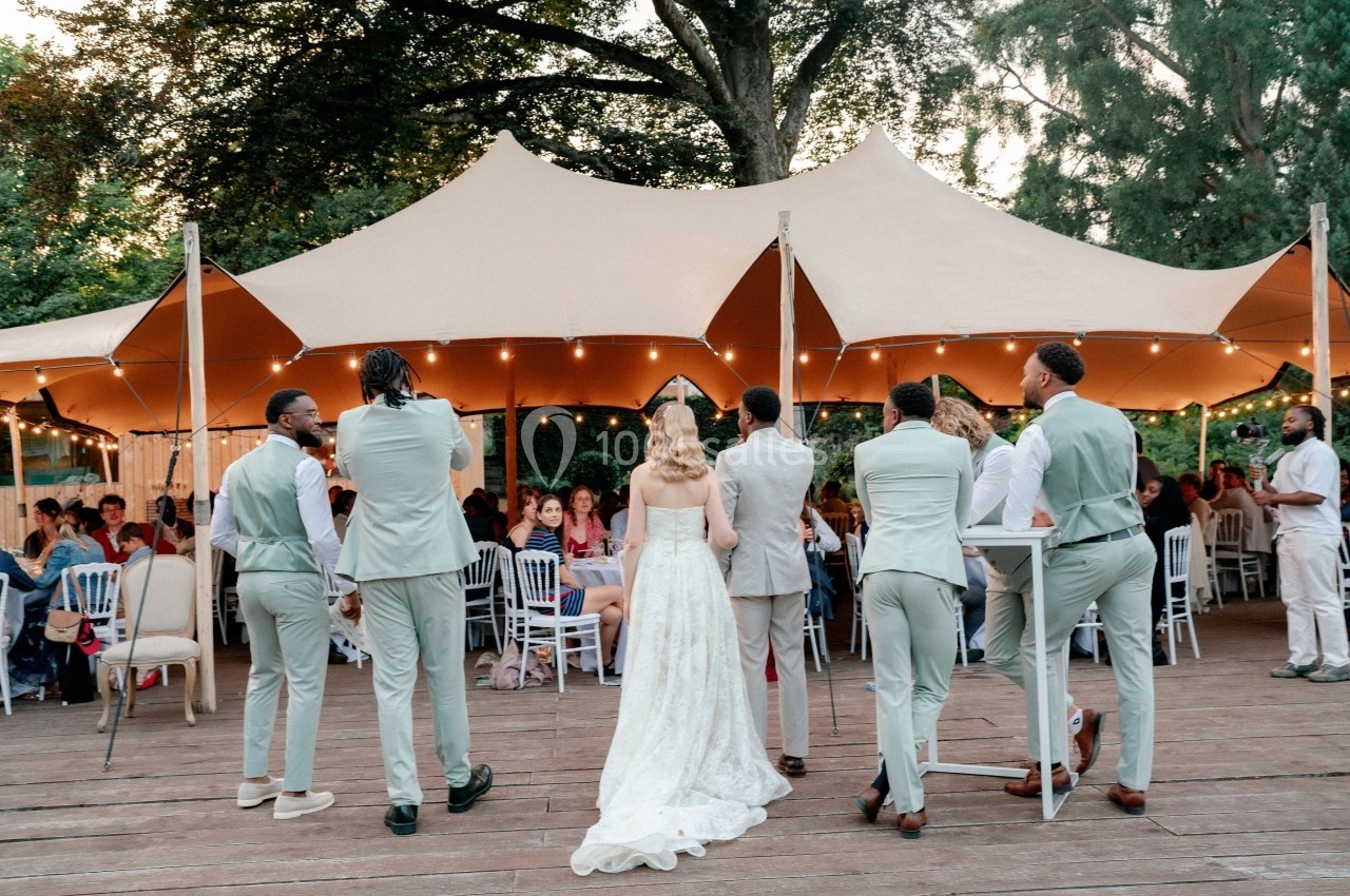 Un groupe de personnes en tenue de mariage se tient devant une tente éclairée lors d'une réception en plein air.