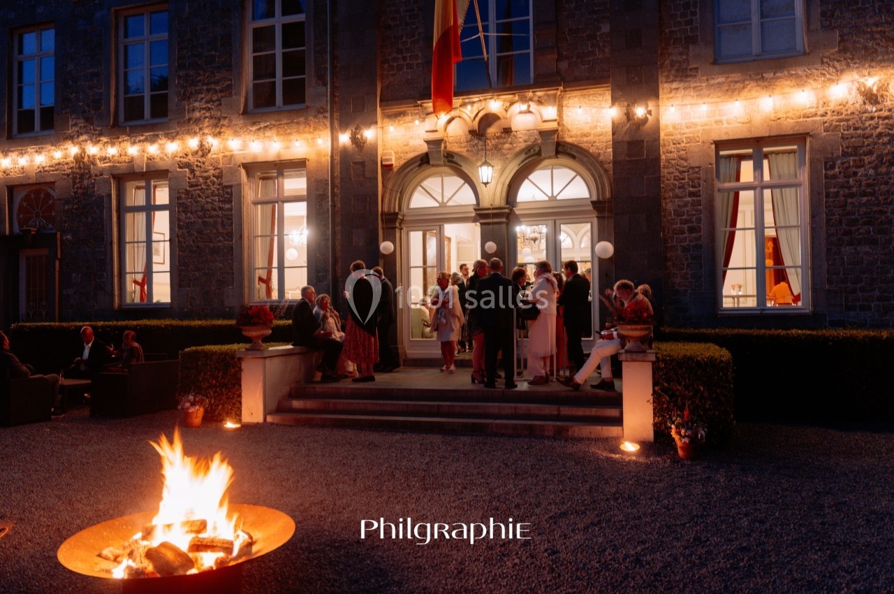 Groupe de personnes discutant devant une entrée éclairée d'un bâtiment en soirée, avec un feu de camp au premier plan.