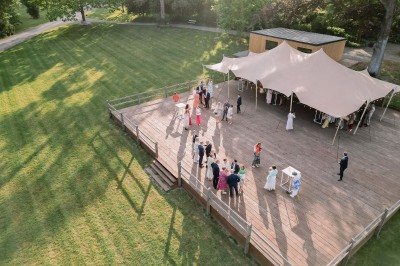 Groupe de personnes rassemblées sous une tente beige sur une terrasse en bois, dans un jardin arboré.