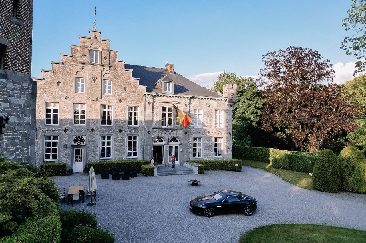 Façade d'un bâtiment historique avec un drapeau belge, une cour gravillonnée et une voiture noire stationnée.