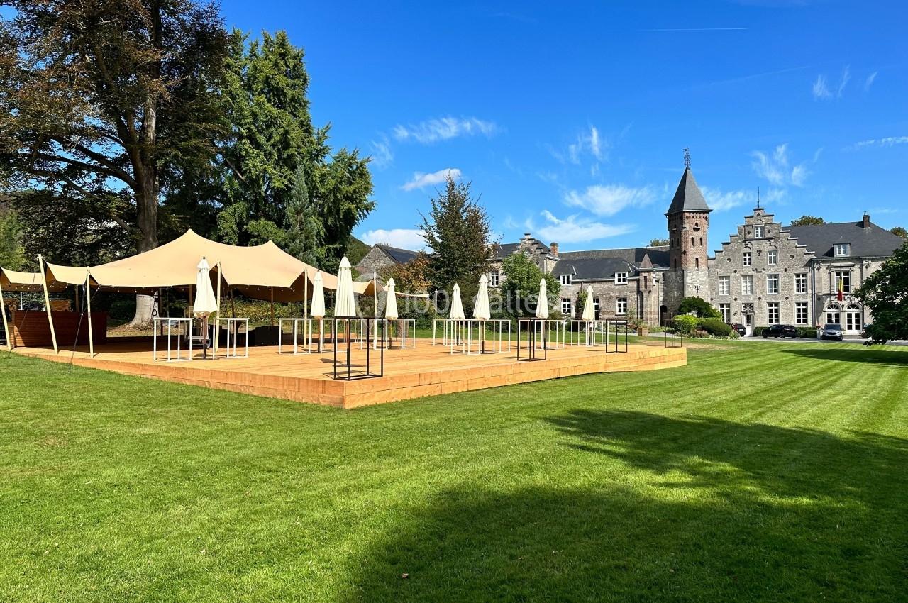 Terrasse en bois avec tables et parasols sur une pelouse, près d'un bâtiment historique sous un ciel bleu.