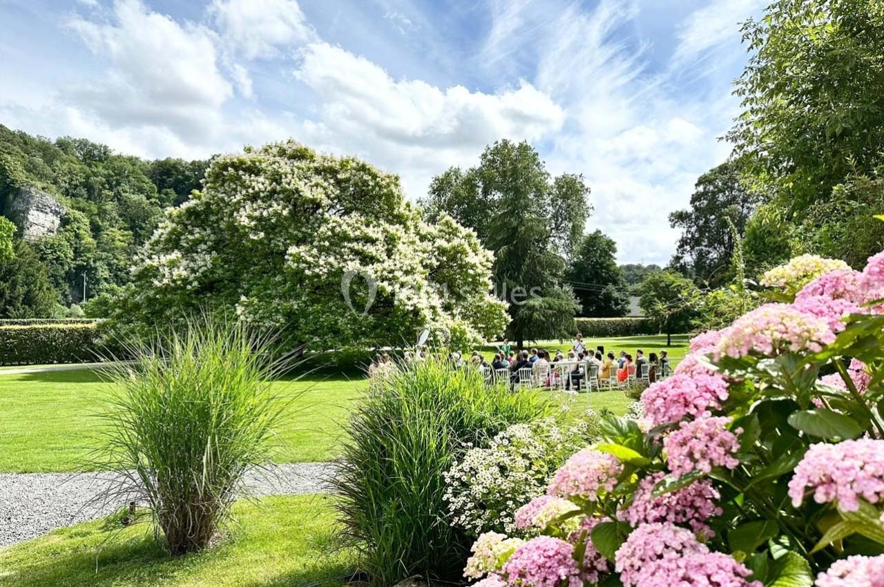 Un jardin verdoyant avec des fleurs roses au premier plan et un groupe de personnes assises à l'extérieur.
