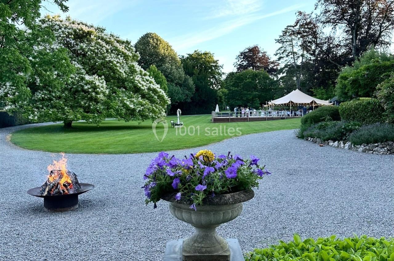 Allée gravillonnée bordée de verdure, avec un brasero, une jardinière fleurie et une tente au fond dans un parc.