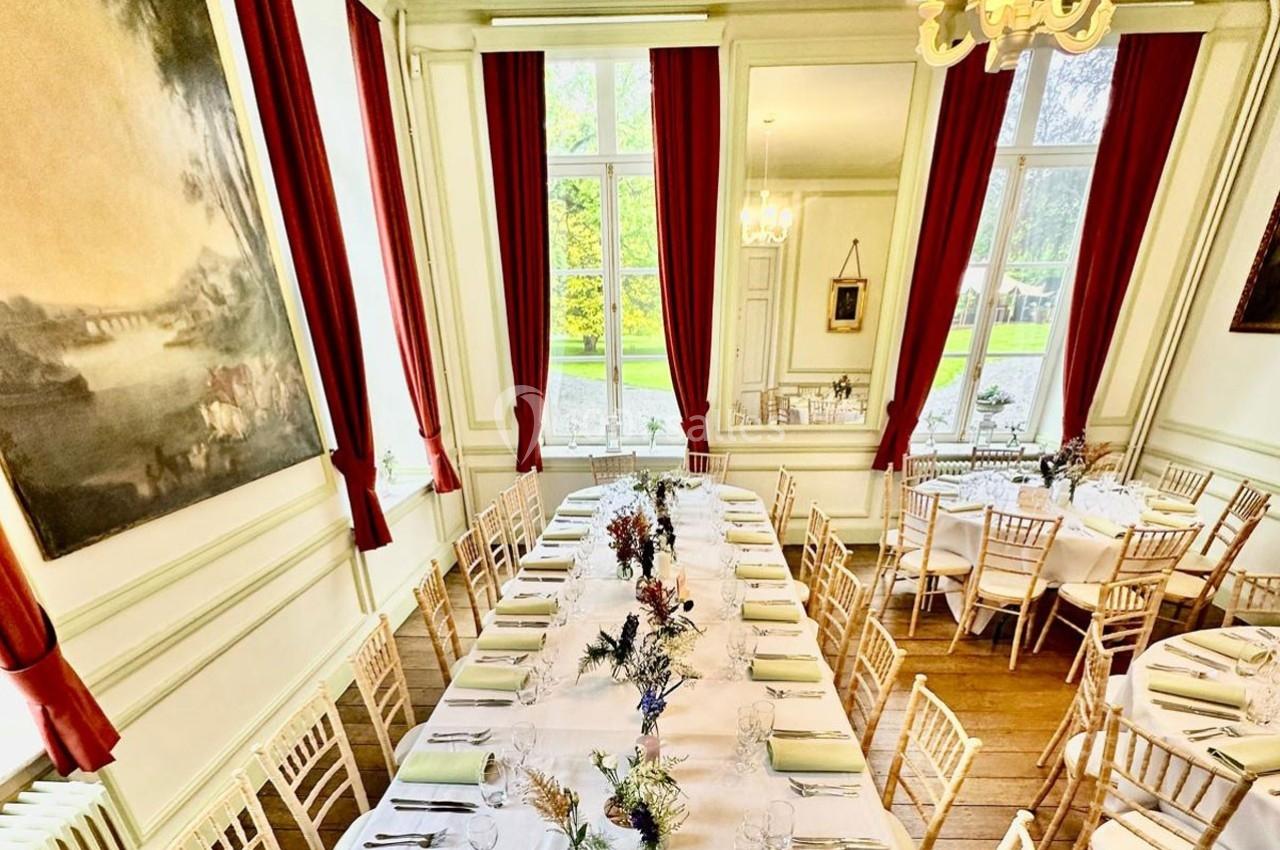 Salle lumineuse avec de grandes fenêtres, tables dressées pour un repas, chaises en bois clair et rideaux rouges.