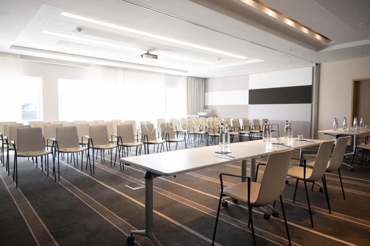 Salle de réunion lumineuse avec des rangées de chaises et des tables équipées de bouteilles d'eau et de verres.