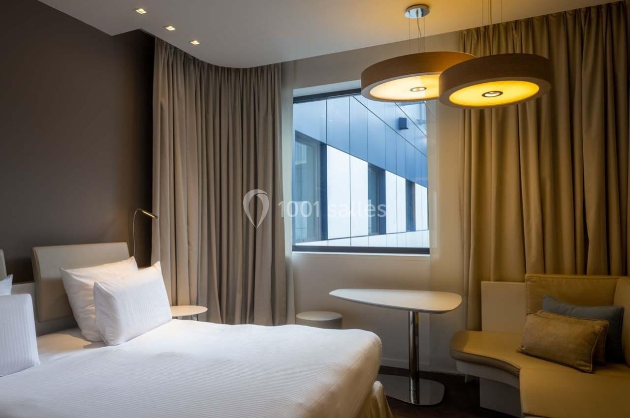 Chambre d'hôtel moderne avec lit double, table, fauteuil, grandes fenêtres et rideaux beiges.