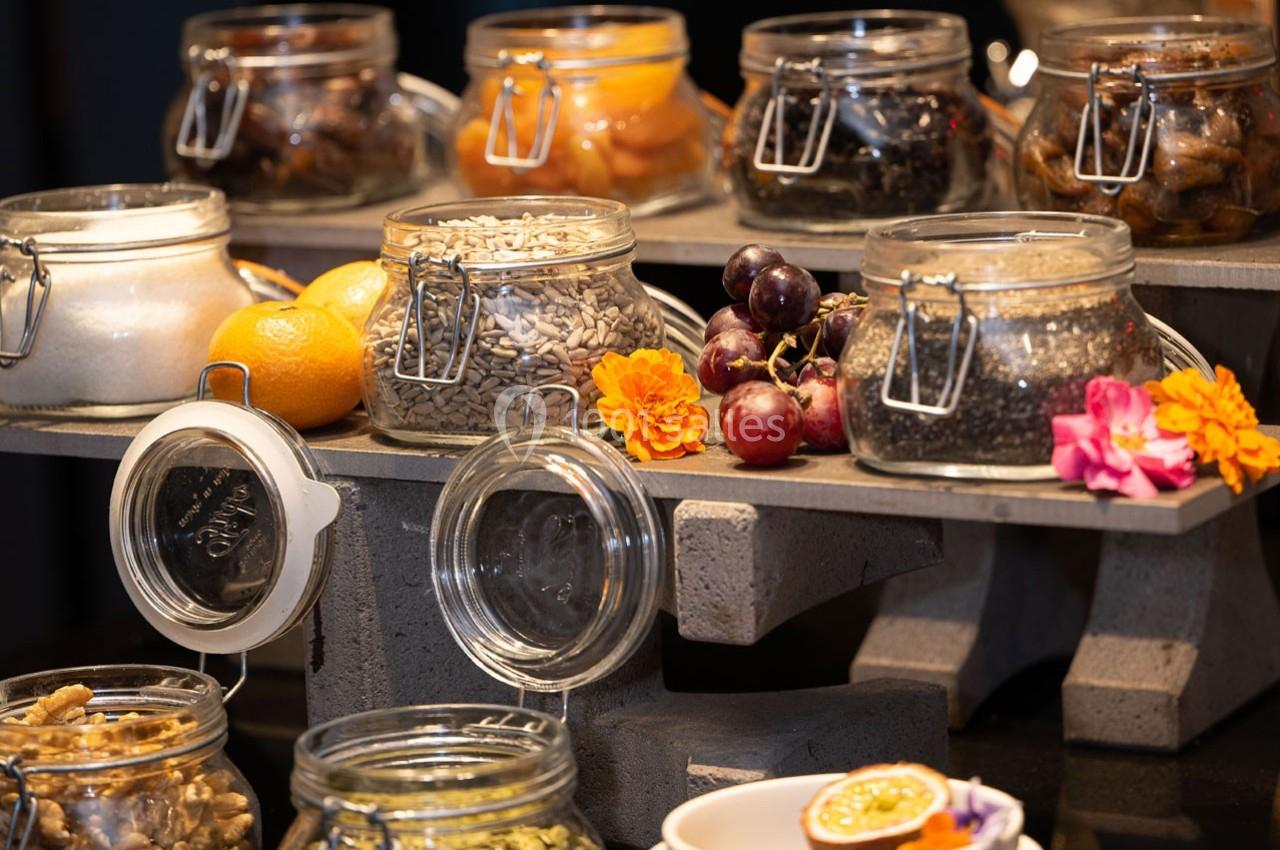 Bocaux en verre contenant graines, fruits secs et confits, disposés sur un présentoir en pierre avec décorations florales.