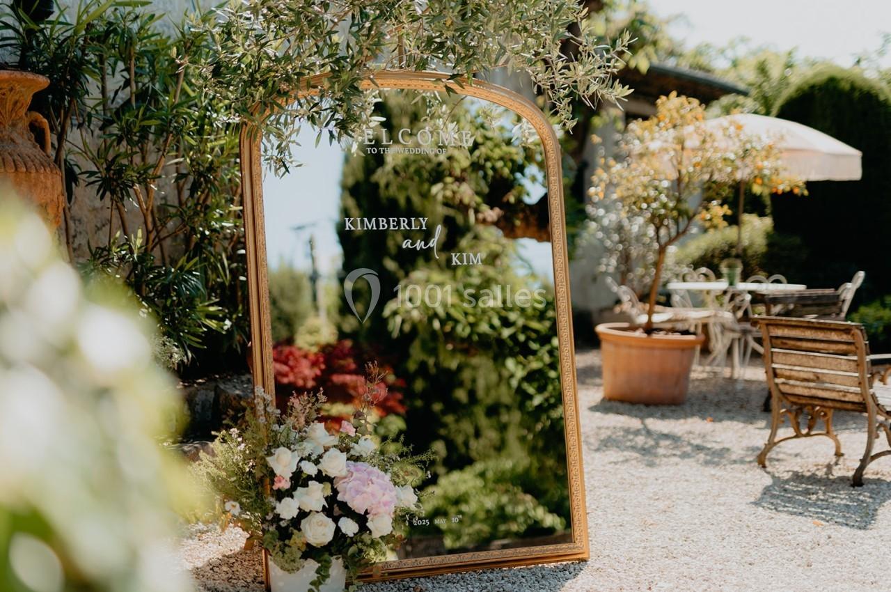 Grand miroir doré avec message de bienvenue gravé, entouré de plantes et fleurs, dans un jardin ensoleillé.