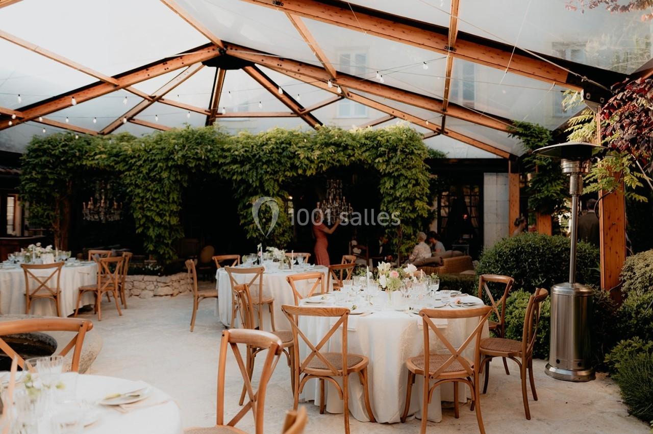 Salle de réception en extérieur avec tables dressées, chaises en bois et toit transparent entouré de verdure.