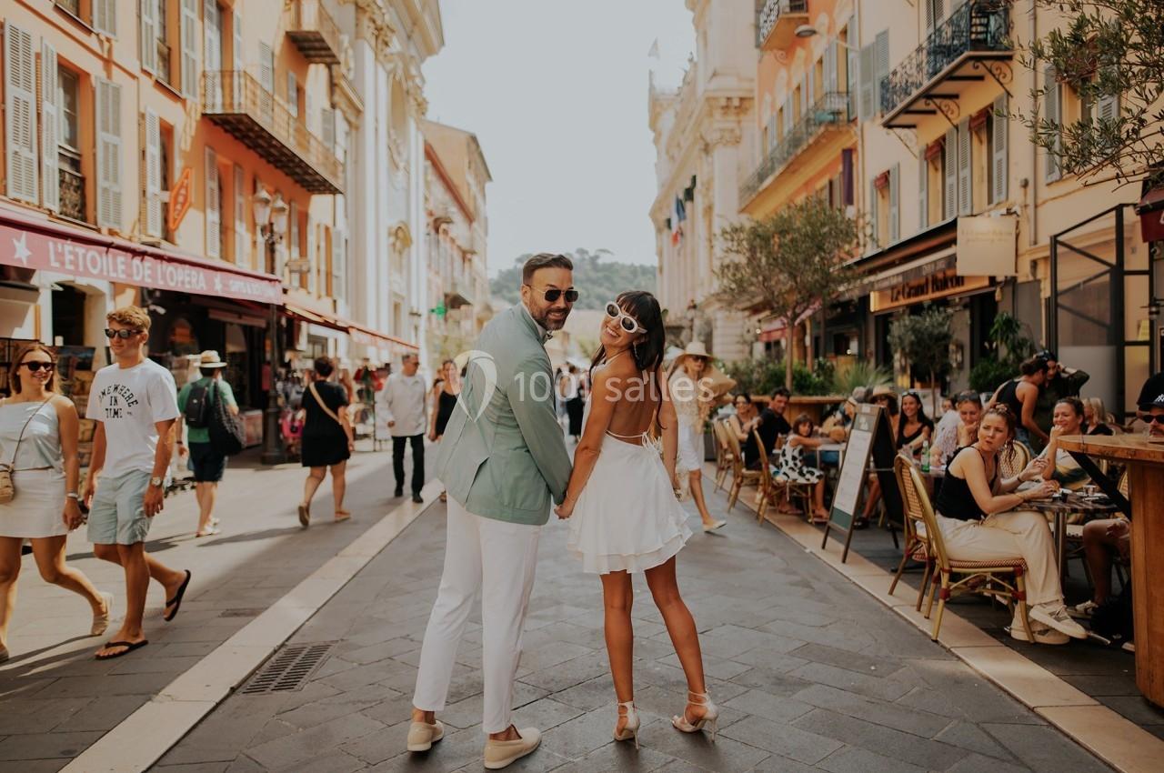 Un couple souriant se tient la main dans une rue animée bordée de cafés et de bâtiments colorés.