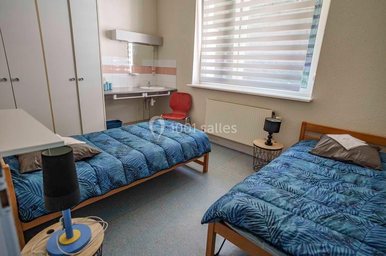 Deux lits simples avec couvre-lits bleus, une lampe de chevet, un bureau et un lavabo dans une chambre lumineuse.